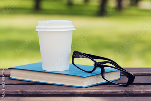 Book, eyeglasses and paper cup with coffee on a bench in park in a sunny day, reading in the summer, education, textbook, back to school concept