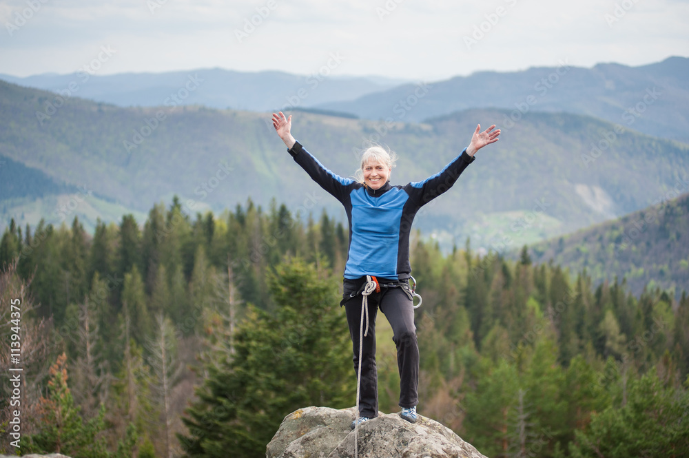 Smiling climber woman in a blue jacket with hands up and with climbing equipment standing on top of mountain on the blurred background of forest valley.