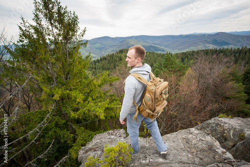 Profile of man with a backpack standing on the edge of a rock and looking into the distance on the green forest and nice mountains. View from above. Wide angle lens © anatoliy_gleb