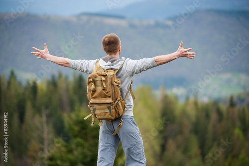 Man with brown backpack standing back to the camera, his arms out to the sides and enjoying picturesque view of on the green forest and nice mountains