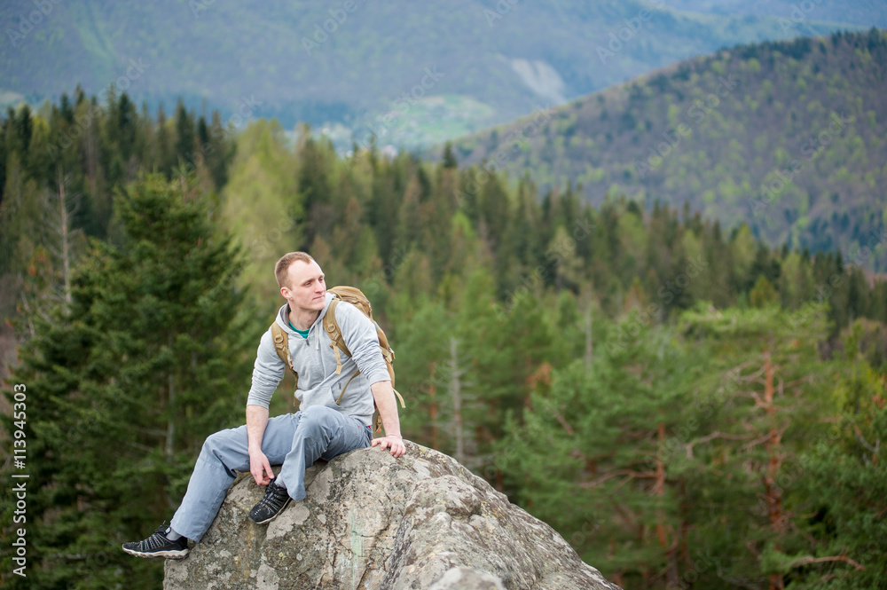 Naklejka premium Athlete man with a brown backpack sitting on the peak of rock and looking down on the blurred background of forest valley and hills. Spring nature