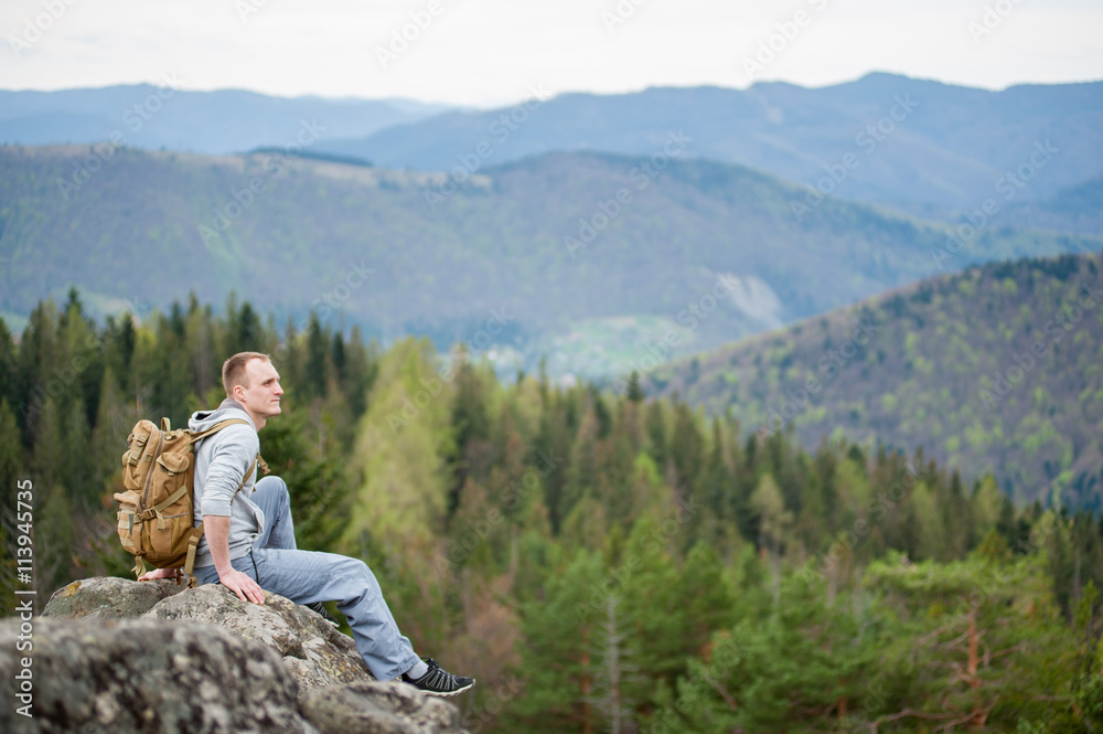 Young tourist man with a backpack sitting on the edge of a rock with an astonishing blurred view on forest valley and hills Young tourist man with a backpack sitting on the edge of a rock with an astonishing blurred view on forest valley and hills