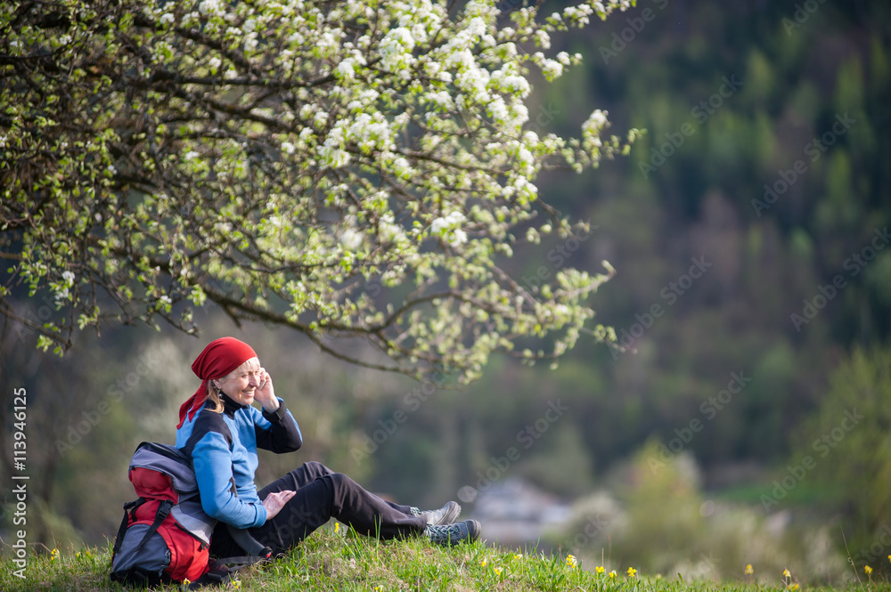 Happy female with a backpack relax under blooming tree on top of hill with yellow wildflowers, green grass and talking on the mobile phone with forest valleys as blurred background. Happy female with a backpack relax under blooming tree on top of hill with yellow wildflowers, green grass and talking on the mobile phone with forest valleys as blurred background.