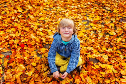 Autumn portrait of a cute little boy of 4 years old  playing with yellow leaves in the park  wearing blue jacket and yellow trousers