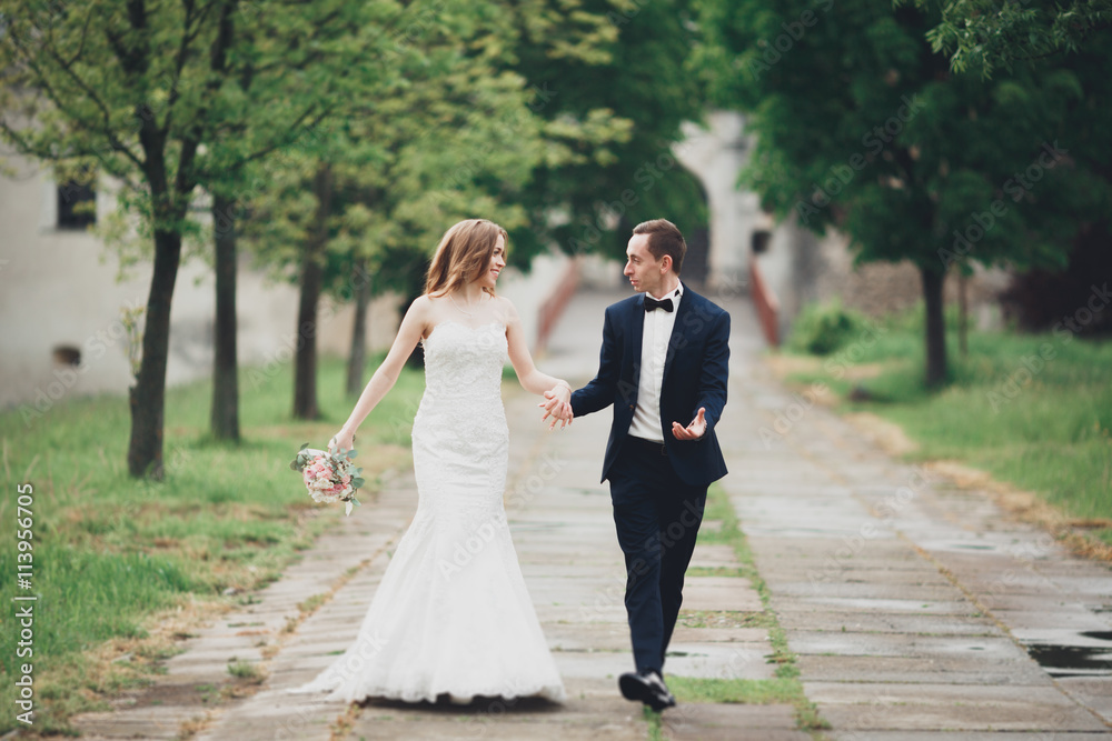 Beautiful couple of happy stylish newlyweds walking in the park on their wedding day with bouquet Beautiful couple of happy stylish newlyweds walking in the park on their wedding day with bouquet