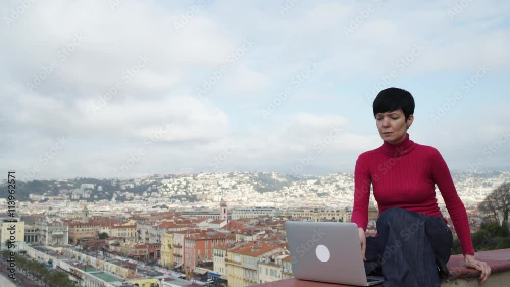 Female typing on the laptop looking on the city view. Nice town Cote d'Azur. Professional businesswoman at holiday vacation. Attractive girl with short hair wearing in casual red pullover and blue Female typing on the laptop looking on the city view. Nice town Cote d'Azur. Professional businesswoman at holiday vacation. Attractive girl with short hair wearing in casual red pullover and blue