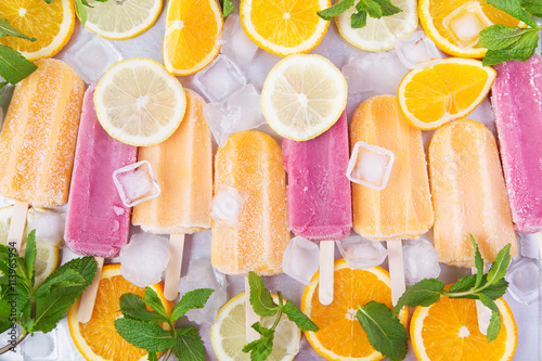 Frosty strawberry and orange ice cream popsicles in a row on a steel tray with mint and ice background