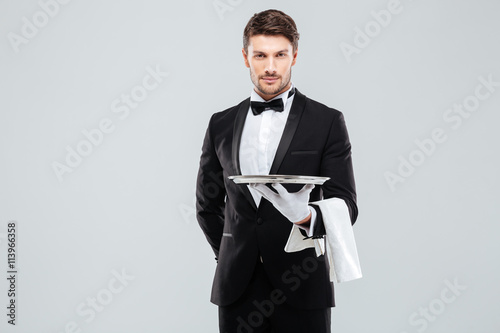 Handsome yong waiter in tuxedo and gloves holding empty tray photo