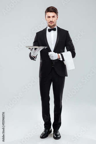 Confident young waiter in tuxedo standing and holding tray photo