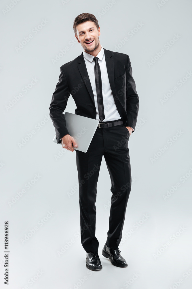 Cheerful handsome young businessman standing and holding laptop Cheerful handsome young businessman standing and holding laptop
