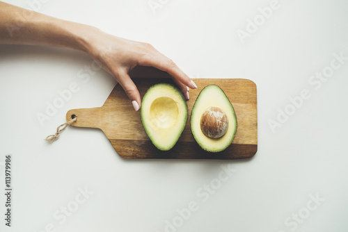 Close-up image of female hand holding fresh avocado on wooden board in the white background, healthy lifestyle concept