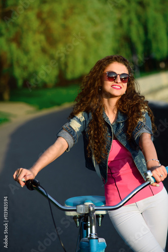 Beautiful girl with curly hair wearing sunglasses riding on a bicycle in the park in the summer in the city