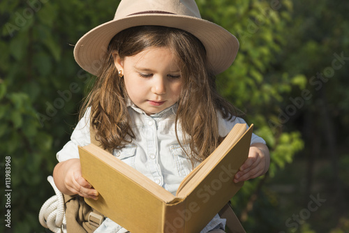 Adorable little girl in a safari hat and explorer clothes reading old book sitting in a wooden suitcase with backpack and safty roap outdoor photo