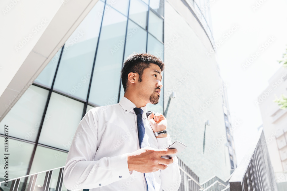 Japanese businessman in suit and tie checking smartphone in front of a modern office building Japanese businessman in suit and tie checking smartphone in front of a modern office building