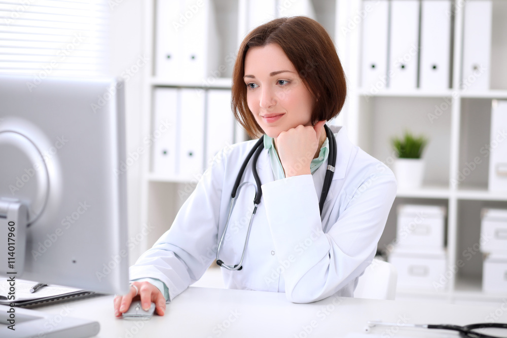 Young brunette female doctor sitting at a desk and working on the computer at the hospital office. Health care, insurance and help concept. Physician ready to examine patient Young brunette female doctor sitting at a desk and working on the computer at the hospital office. Health care, insurance and help concept. Physician ready to examine patient