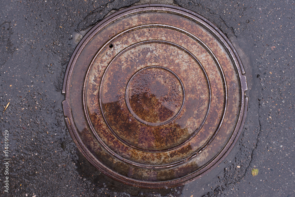 Closeup photo of Old Sewer rust manhole cover on the urban asphalt road. Rain scene.