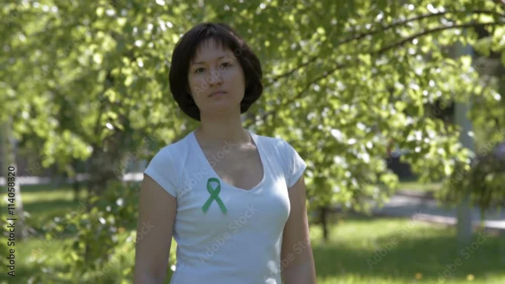 Woman pinning green ribbon on white t-shirt, trees in background. Kidney cancer, glaucoma, organ donation, depression, environmental protection awareness Woman pinning green ribbon on white t-shirt, trees in background. Kidney cancer, glaucoma, organ donation, depression, environmental protection awareness