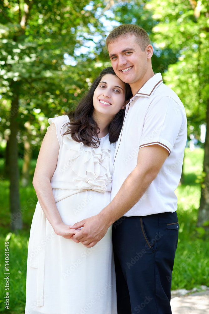 pregnant woman with husband posing in the city park, family portrait, summer season, green grass and trees pregnant woman with husband posing in the city park, family portrait, summer season, green grass and trees