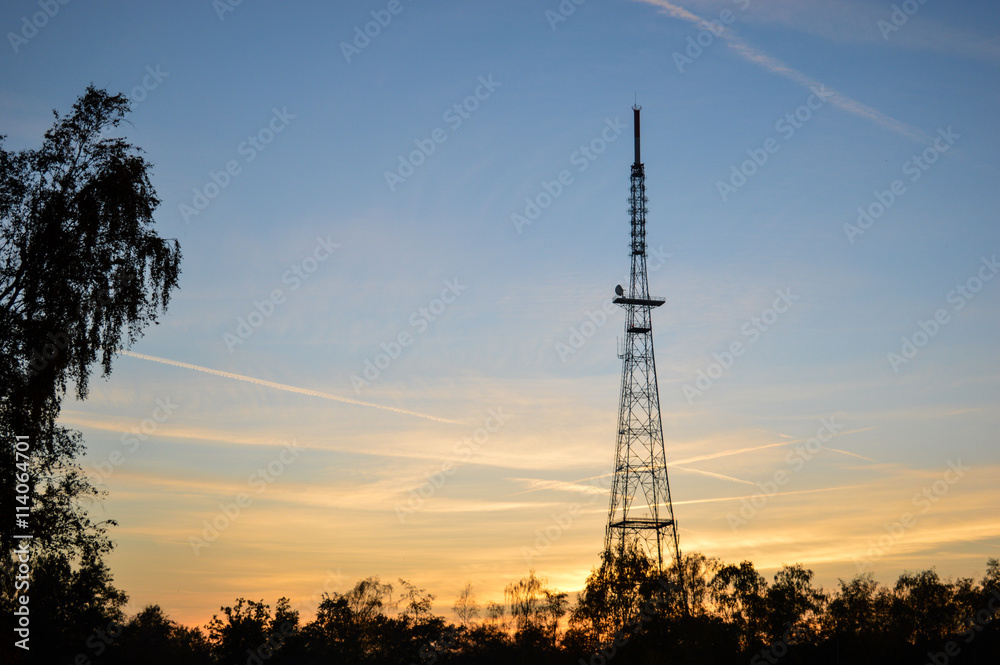 Steel radio tower with trees on the left and bottom at sunset coloring the sky blue and orange