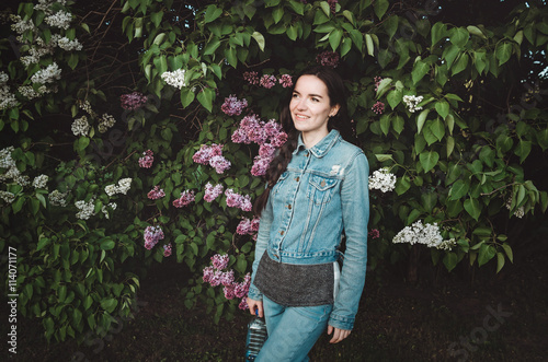 Portrait of a beautiful smiling  young woman outdoor with blossom purple lilac flowers in  spring garden. Attractive female in casual jeans clothes  holding  bottle  water.  concept  nature and care.
