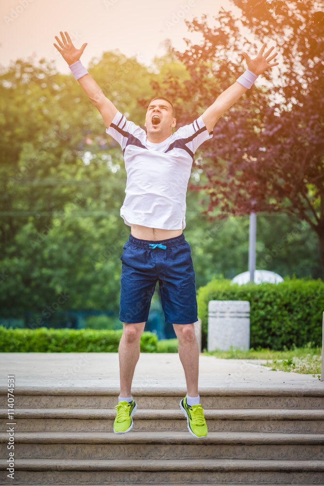 Fototapeta premium Happy jogger jumping on the stairs in the park with raised hands. Another successful training is finished. Sport and fitness success concept. 