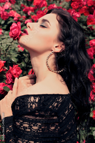 fashion outdoor photo of gorgeous sensual woman with dark hair and bright makeup, in elegant black lace dress posing beside roses at summer garden 