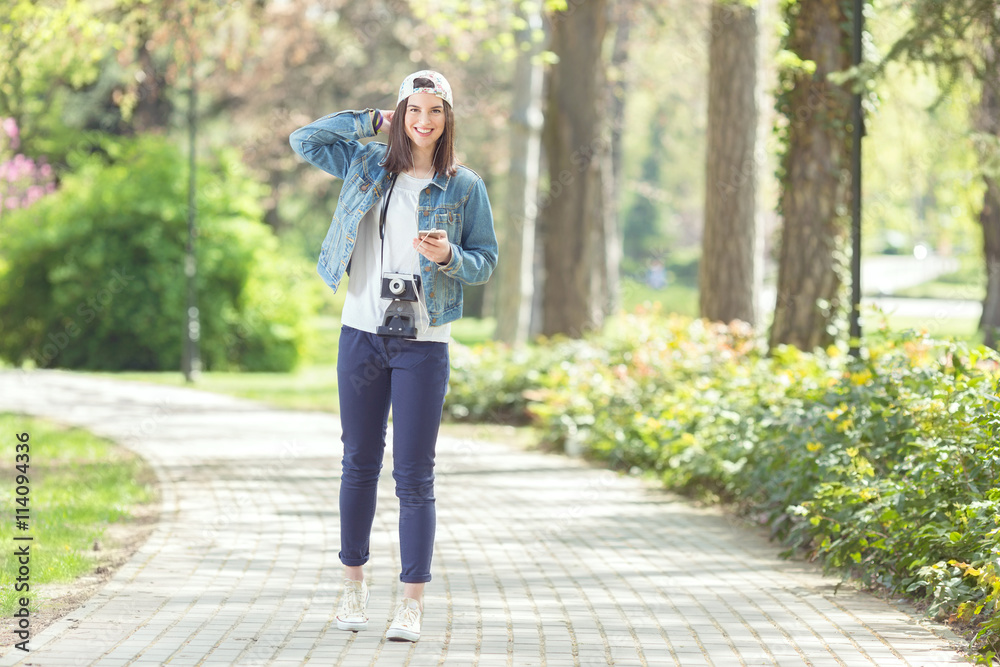 Happy teenage girl is walking in a park and holding mobile phone in her hand, and camera around her neck. She is listening to music on headphones. Happy teenage girl is walking in a park and holding mobile phone in her hand, and camera around her neck. She is listening to music on headphones.