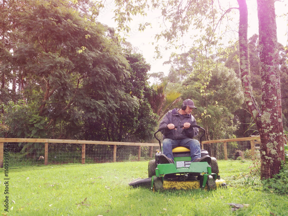 A baby boomer is mowing grass on a rural property on a ride on lawn mower. Filtered image for a vintage retro look with lens flare and grain.
