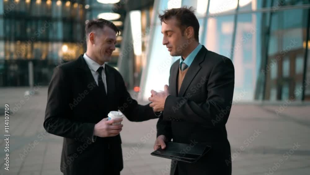 Young successful european businessman treat coffee cup his friend with tablet PC. Smiling conversation. Men going work together. Glass business-centre building at the background. Teal and Orange style Young successful european businessman treat coffee cup his friend with tablet PC. Smiling conversation. Men going work together. Glass business-centre building at the background. Teal and Orange style