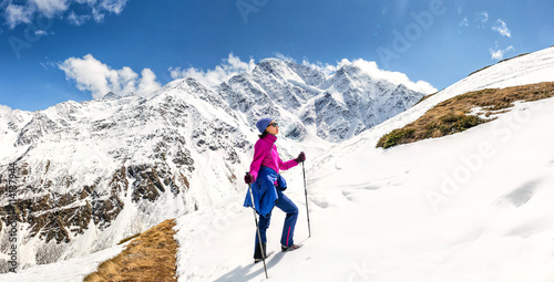 Sunny mountain slope covered with snow and woman climbing with trekking poles. Outdoor among mountain peaks concept