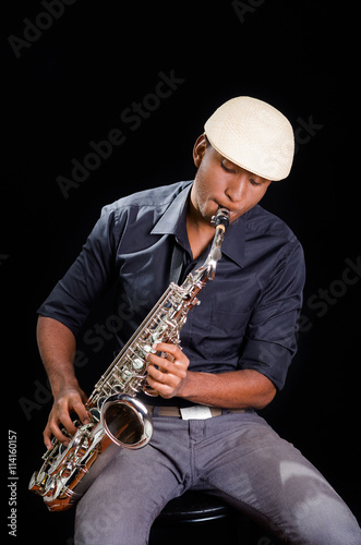 With white hat a black man is sitting and playing his saxophone  dark background  nice music