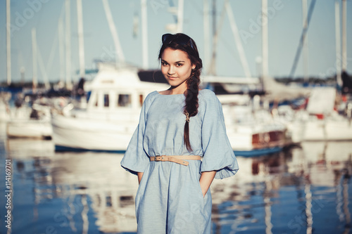 Portrait of beautiful white Caucasian brunette woman with tanned skin in blue dress, by seashore lakeshore with blurry yachts boats on background on water, sunset on summer day, lifestyle concept