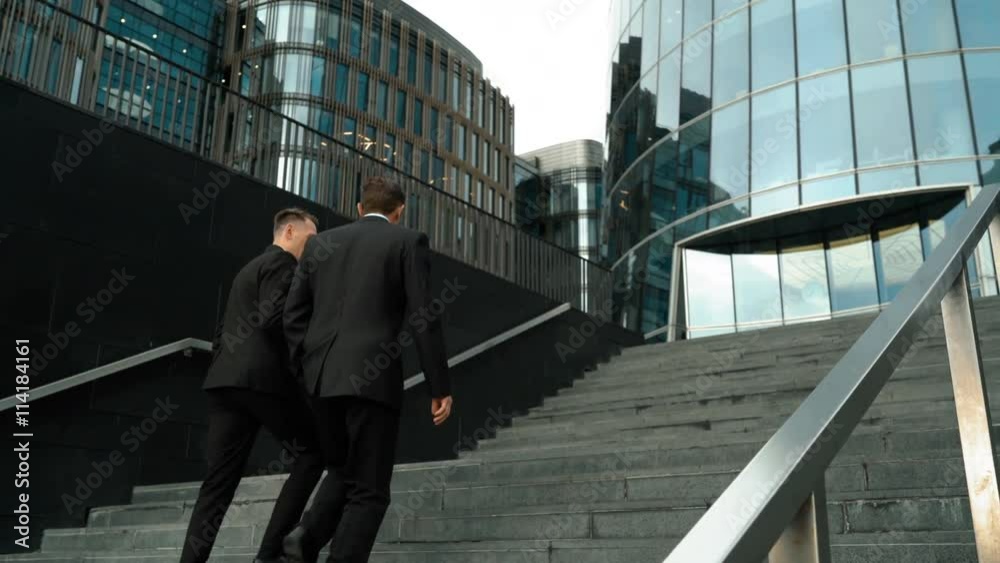 Two young friendly businessmen communicating with carefree morning conversation. They climbing the stairs. Glass business centre building at the background. Teal and Orange style. Wide UltraHD