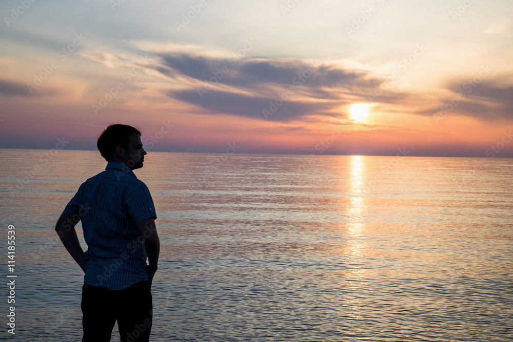Fototapeta premium Young man standing at the beach in front of amazing sea view at sunset or sunrise and thinking about his future. Rear view