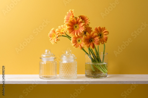 黄色の背景とオレンジ色のガーベラと花瓶とガラス瓶