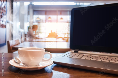 White cup of hot coffee with laptop on wooden table in cafe.
