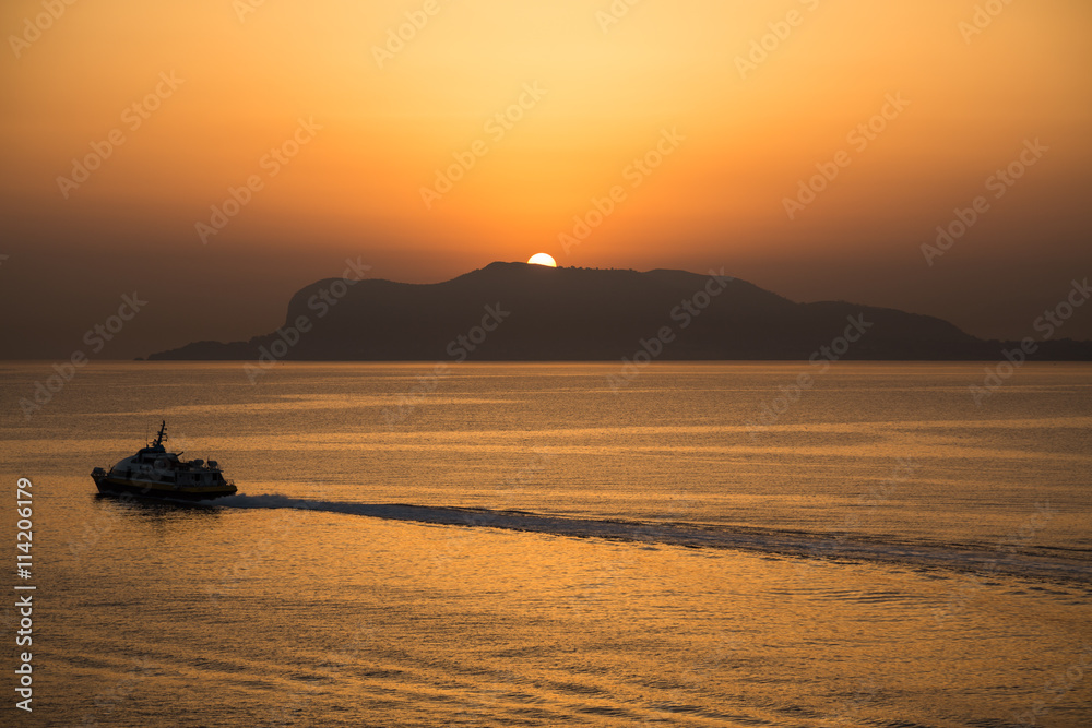 The sun peeks over the mountains and the sea in Sicily, with a silhouette and a wake of a ferry