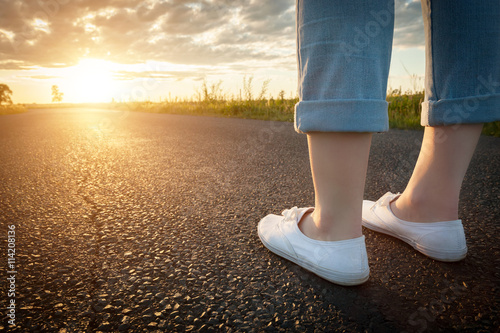 Woman in white sneakers standing on asphalt road towards sun. Travel, freedom concepts.