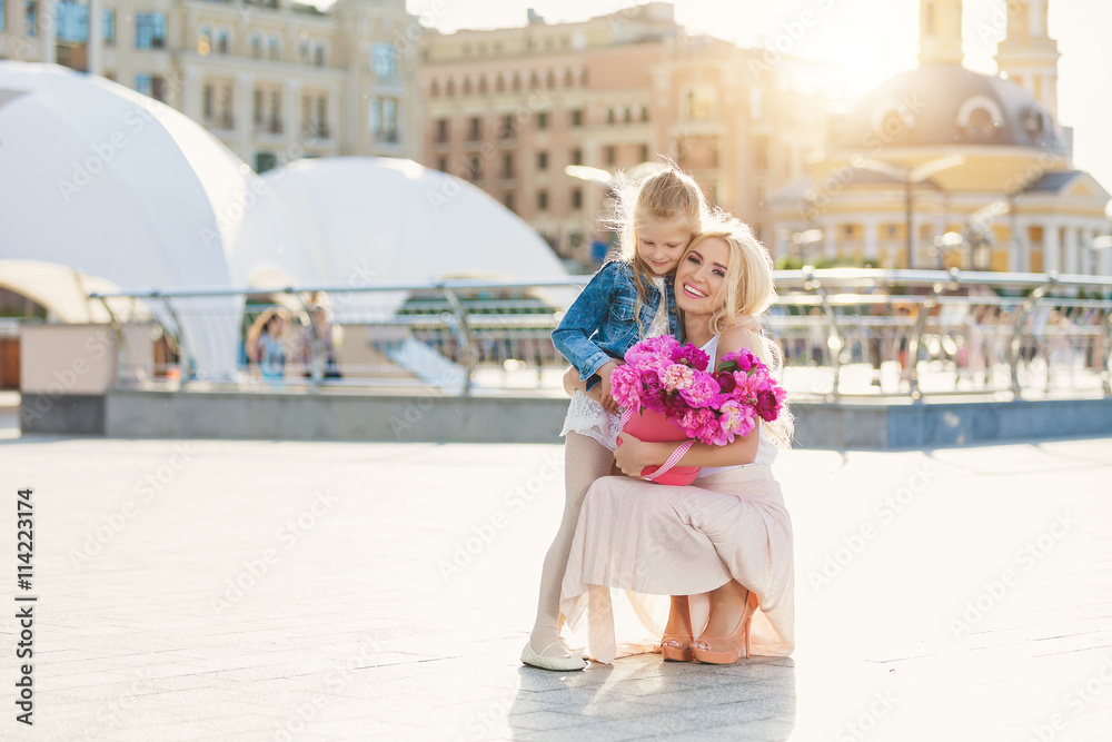 Obraz premium Portrait of young beautiful mother and daughter with box of peonies posing in the city streets. Flower box and gift box. Bouquet of flowers in gift box. Birthday. March 8