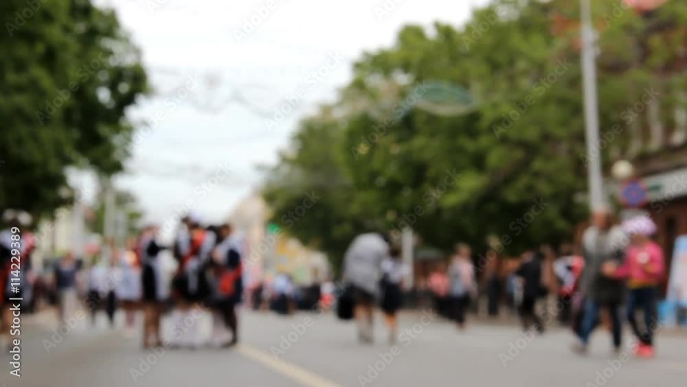 Blurred background. Different walking people. Women with child. Wide street built up by small buildings and green trees. Blurred background. Different walking people. Women with child. Wide street built up by small buildings and green trees.