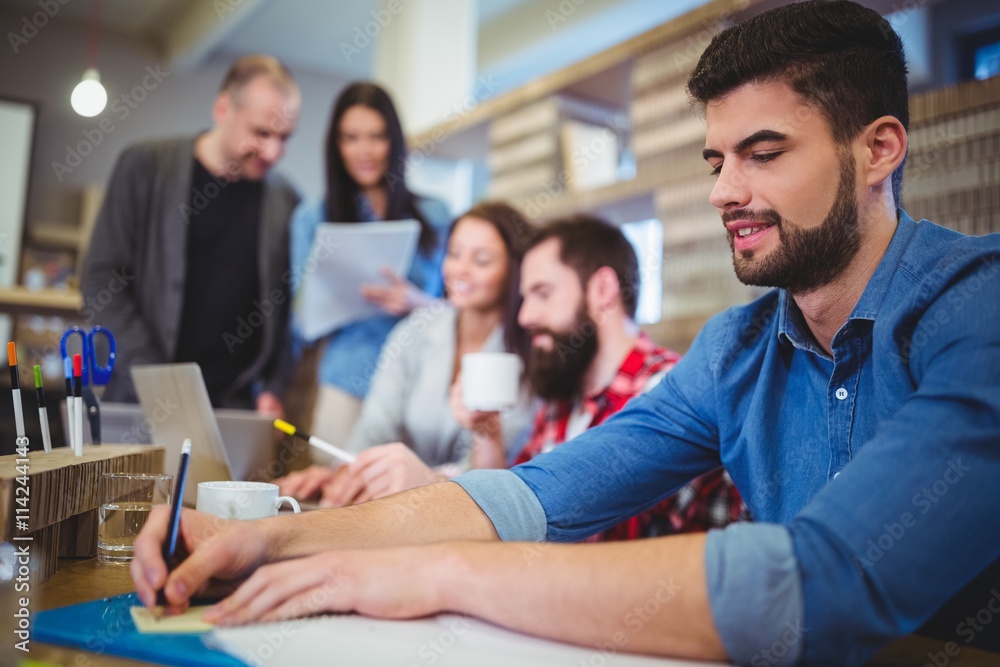 Businessman writing on paper while coworkers in background Businessman writing on paper while coworkers in background
