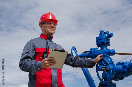 Worker near well head valve holding tablet computer and wearing red helmet in the oilfield. Oil and gas concept. 