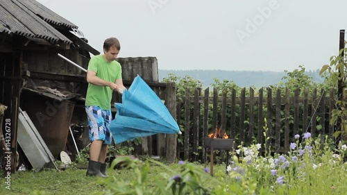 Man with umbrella near the fire for barbecue. The rain is falling, an old building with a fence in the background