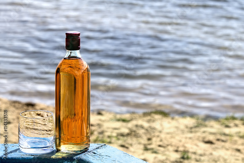 Bottle whiskey and tumbler on shore of lake