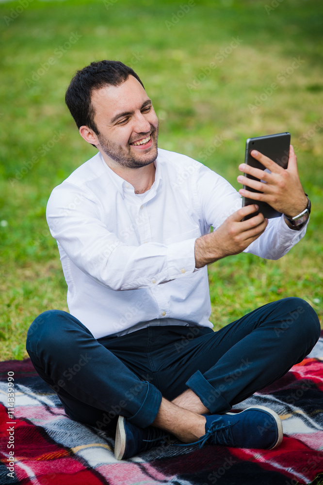 Fototapeta premium Modern young hispanic man taking a selfie at the park, with his tablet.