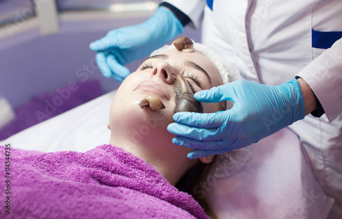 Young woman receiving snail facial massage. Snail on face. Cleaning procedure in spa salon. Blurred background.