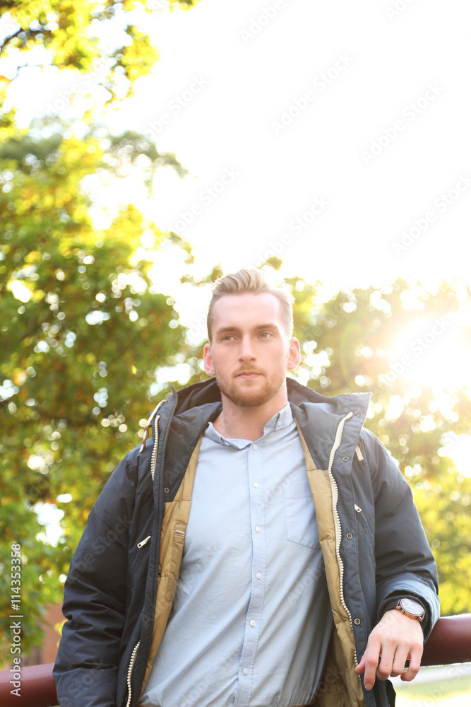 Obraz premium An attractive blonde man standing leaning outdoors on a sunny day with the sun right behind him, lens flare. Looking away from camera.