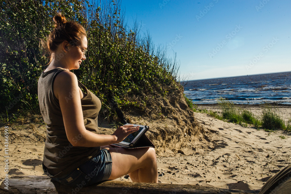 Woman in glasses using digital tablet at sunny sea sand coast. Freelance and rest concept.