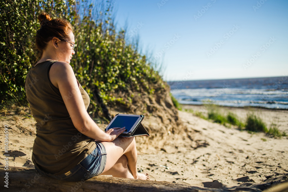 Woman in glasses using digital tablet at sunny sea sand coast. Freelance and rest concept.