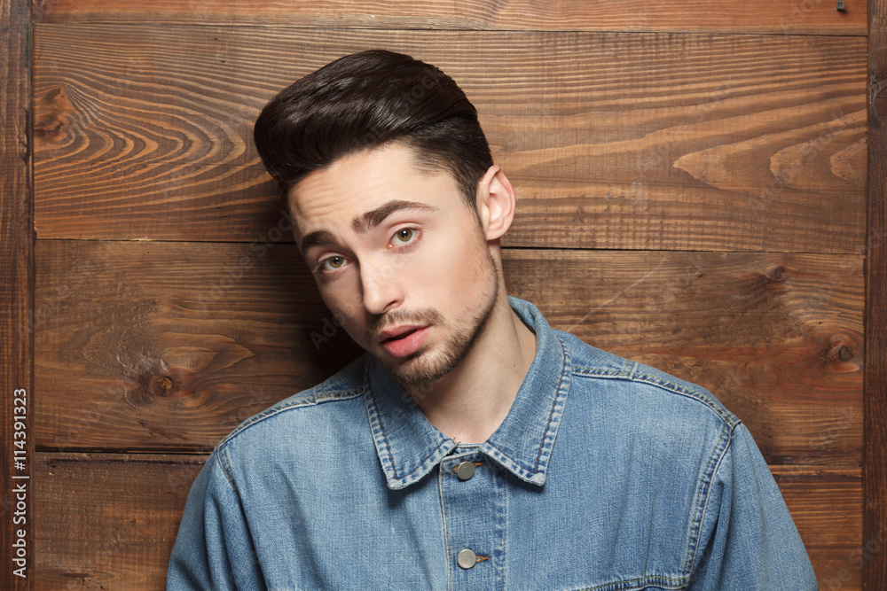 Portrait of handsome model man in jeans shirt posing over wooden background while demonstarting his modern hairstyle in studio. Portrait of handsome model man in jeans shirt posing over wooden background while demonstarting his modern hairstyle in studio.